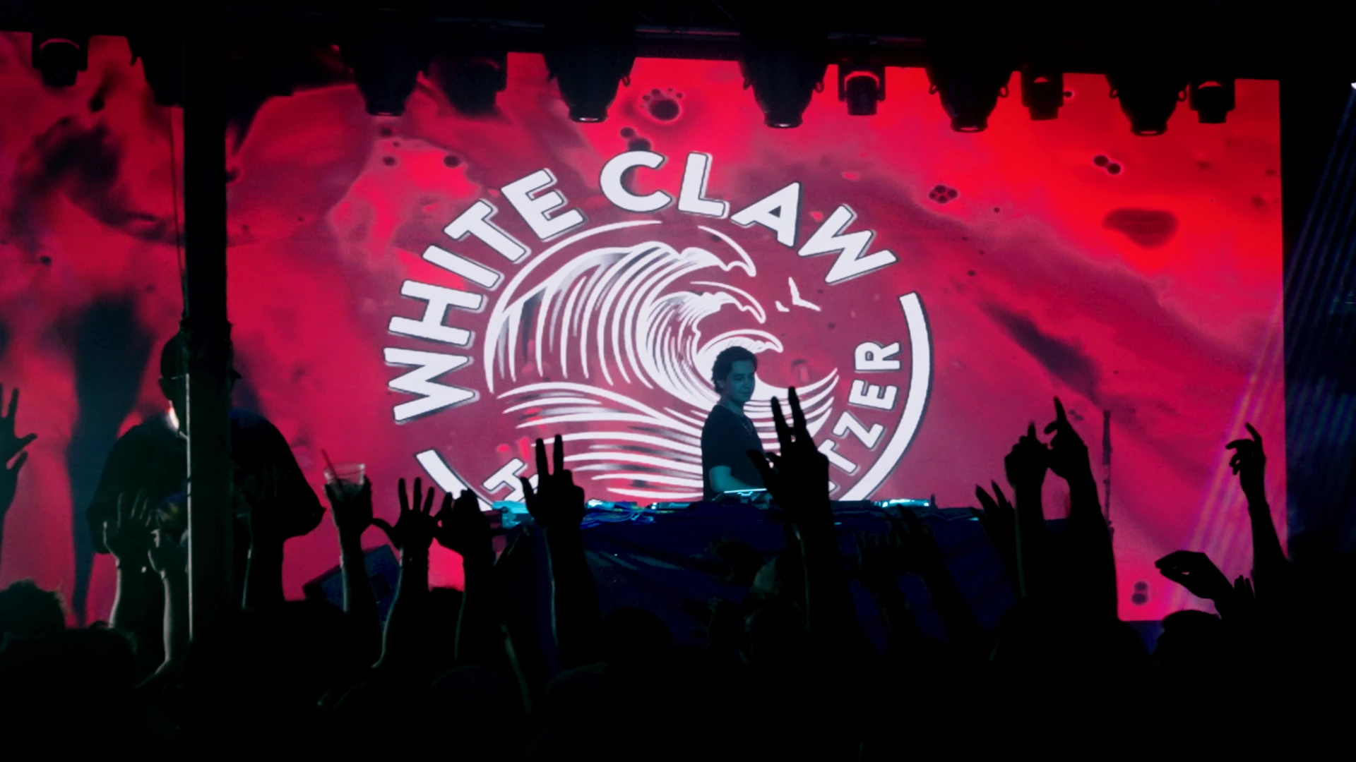 DJ performing in front of a red White Claw backdrop as the crowd raises their hands.