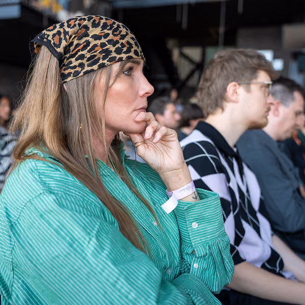 Woman in a green striped shirt and leopard-print headscarf rests her chin on her hand, listening intently among a group of seated people.