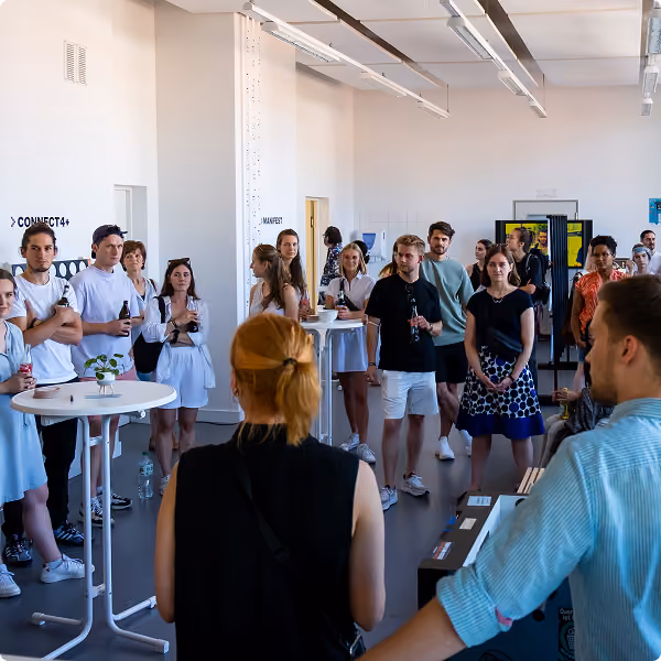 A group of young adults standing in a spacious, brightly lit room, some holding drinks, appearing to listen attentively to a speaker whose back is to the camera.