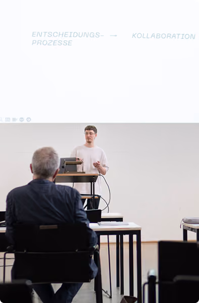 A young man gives a presentation in a seminar room, standing behind a podium with a laptop and microphone, facing an older man seated at a desk with a laptop.
