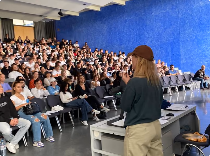 A person wearing a brown cap and green sweater speaks to a large audience seated in rows in a room with a blue wall.