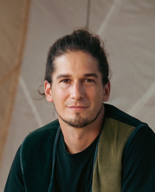 Portrait of a young man (Simon) with light skin and brown hair tied back, wearing a dark shirt and green vest, looking at the camera with a slight smile against a neutral background.