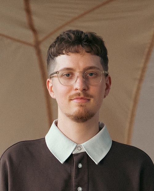 Young man (Luca) with short curly hair, wearing clear glasses and a brown shirt with a large light blue collar, looking directly at the camera with a neutral expression.