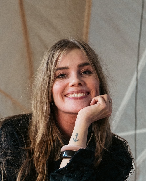 Smiling young woman (Alexandra) with long blond hair resting her chin on her hand, showing a small anchor tattoo on her wrist and wearing a black top.