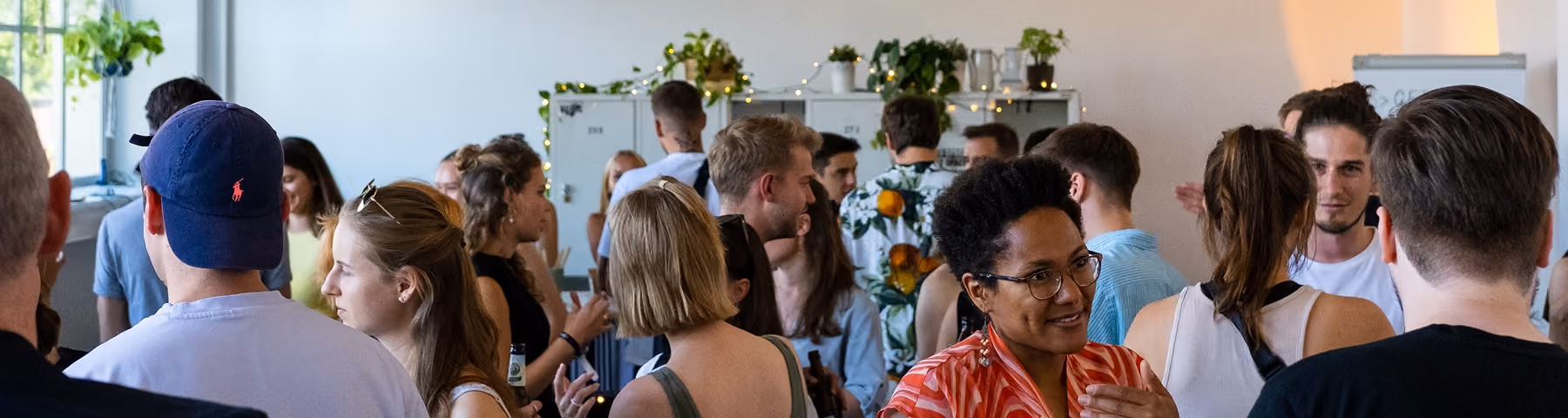 A diverse group of people socializing indoors in a well-lit room with plants and decorations on a shelf in the background.