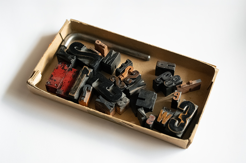 A shallow rectangular cardboard tray holding assorted vintage wooden letterpress blocks in black and reddish colors on a white surface.