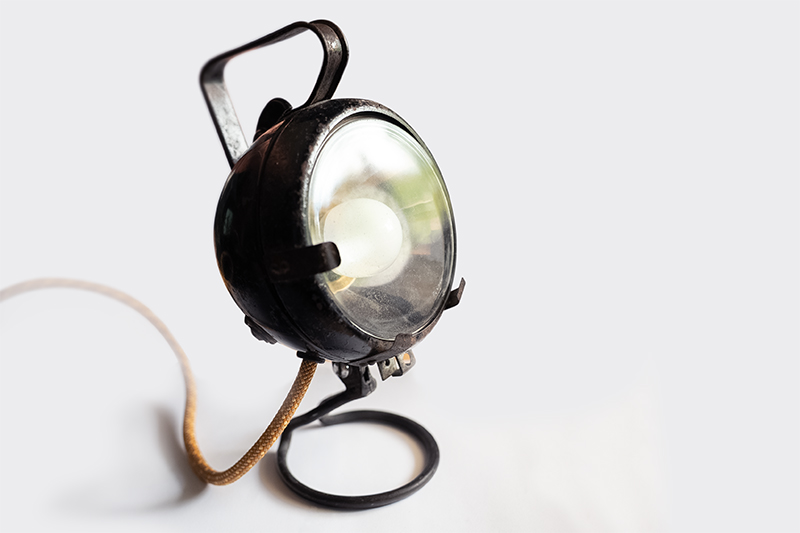 Vintage black metal spotlight lamp with a glass bulb cover on a circular base, connected to a brown cloth-covered cord, against a plain light background.