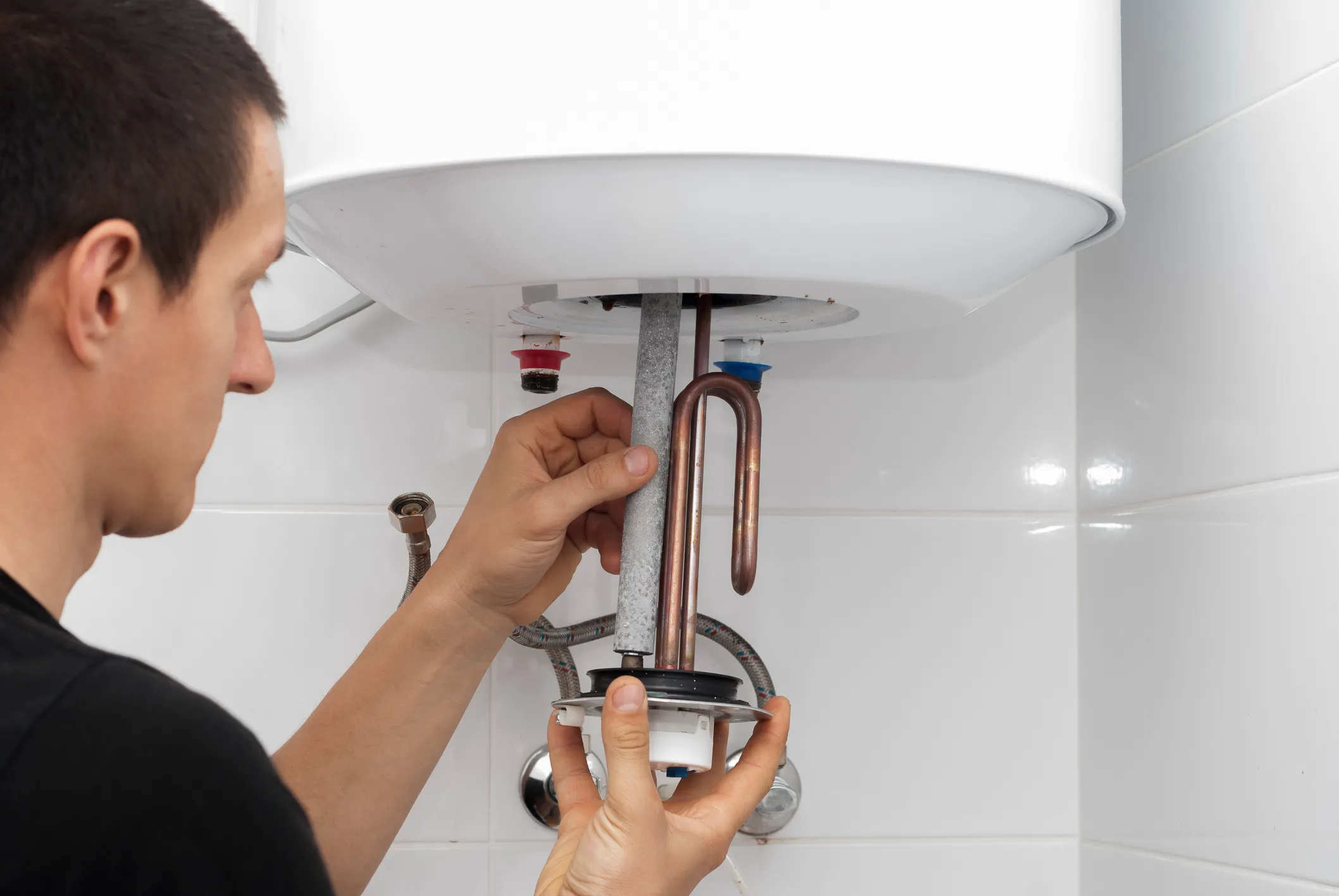 A person's hands are shown removing a heating element and anode rod from the bottom of an electric water heater, revealing the interior.
