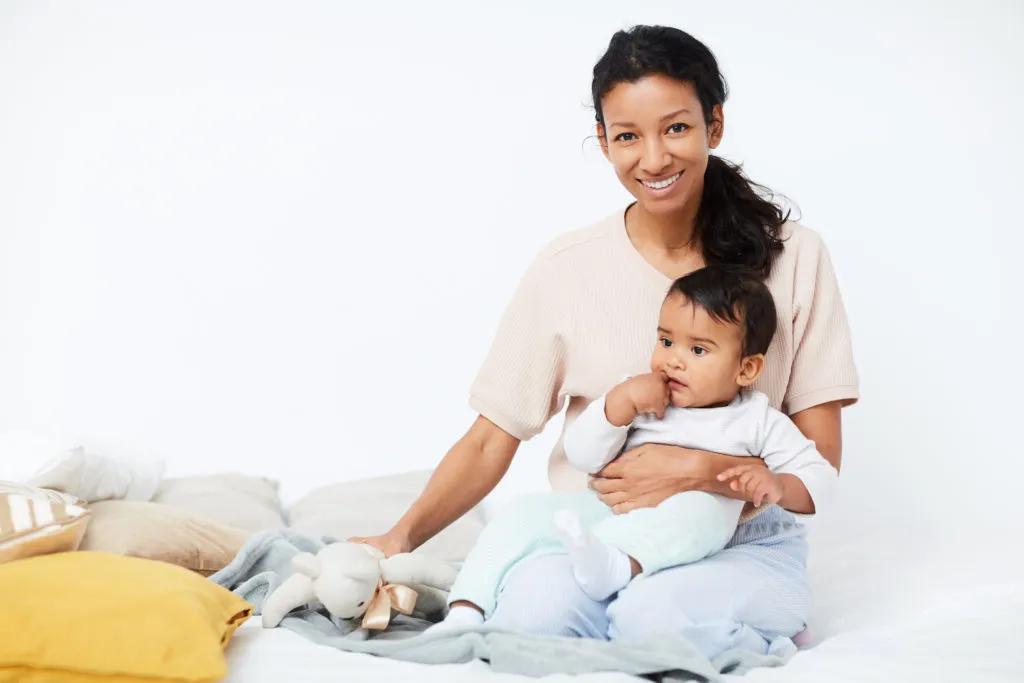  A happy mother is sitting on a bed with her baby. The mother has dark hair tied back in a ponytail and is wearing a light-colored top. She is smiling directly at the camera. She is holding the baby, who is looking away from the camera and has their hand in their mouth. The baby is wearing a white shirt and light green pants. A stuffed bunny is on the bed next to them, and several pillows are scattered around. The background is a simple, white wall.
