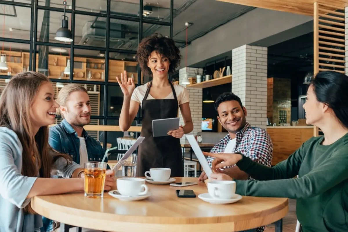  A young waitress with curly hair and a brown apron is smiling and waving to a group of four customers seated at a round wooden table in a cafe. She is holding a tablet, ready to take their order. The customers, two women and two men, are also smiling and engaged in conversation. One woman is pointing at a menu. The cafe has a modern, industrial look with exposed brick and a glass grid wall. A variety of drinks and coffee cups are on the table.
