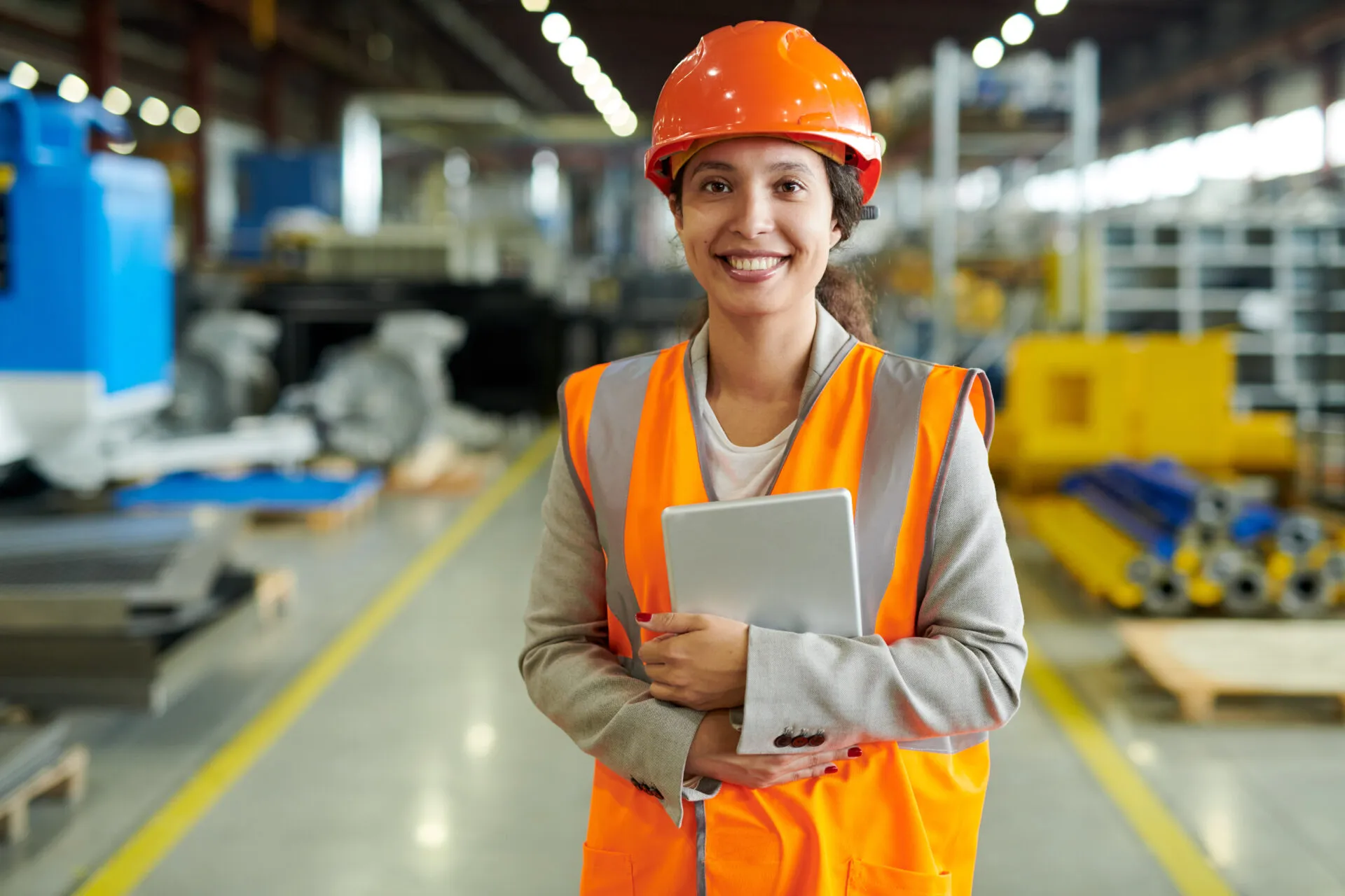A professional-looking female factory worker is smiling and looking at the camera. She is standing in a factory or warehouse aisle with yellow-striped floors and machinery in the background. She is wearing a bright orange safety vest over a gray blazer and a white shirt. She is also wearing a bright orange hard hat and holding a silver tablet. Her dark hair is pulled back, and she has a warm, confident expression.
