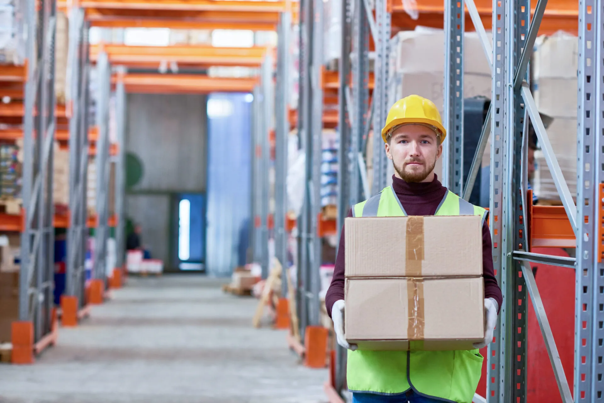  A male warehouse worker is standing in the middle of an aisle, holding two cardboard boxes. He is wearing a yellow hard hat, a burgundy turtleneck, and a bright green safety vest. He is also wearing white work gloves. In the background, there are tall metal shelves filled with various items and a bright light from a doorway at the far end of the aisle. He is looking directly at the camera with a neutral expression.