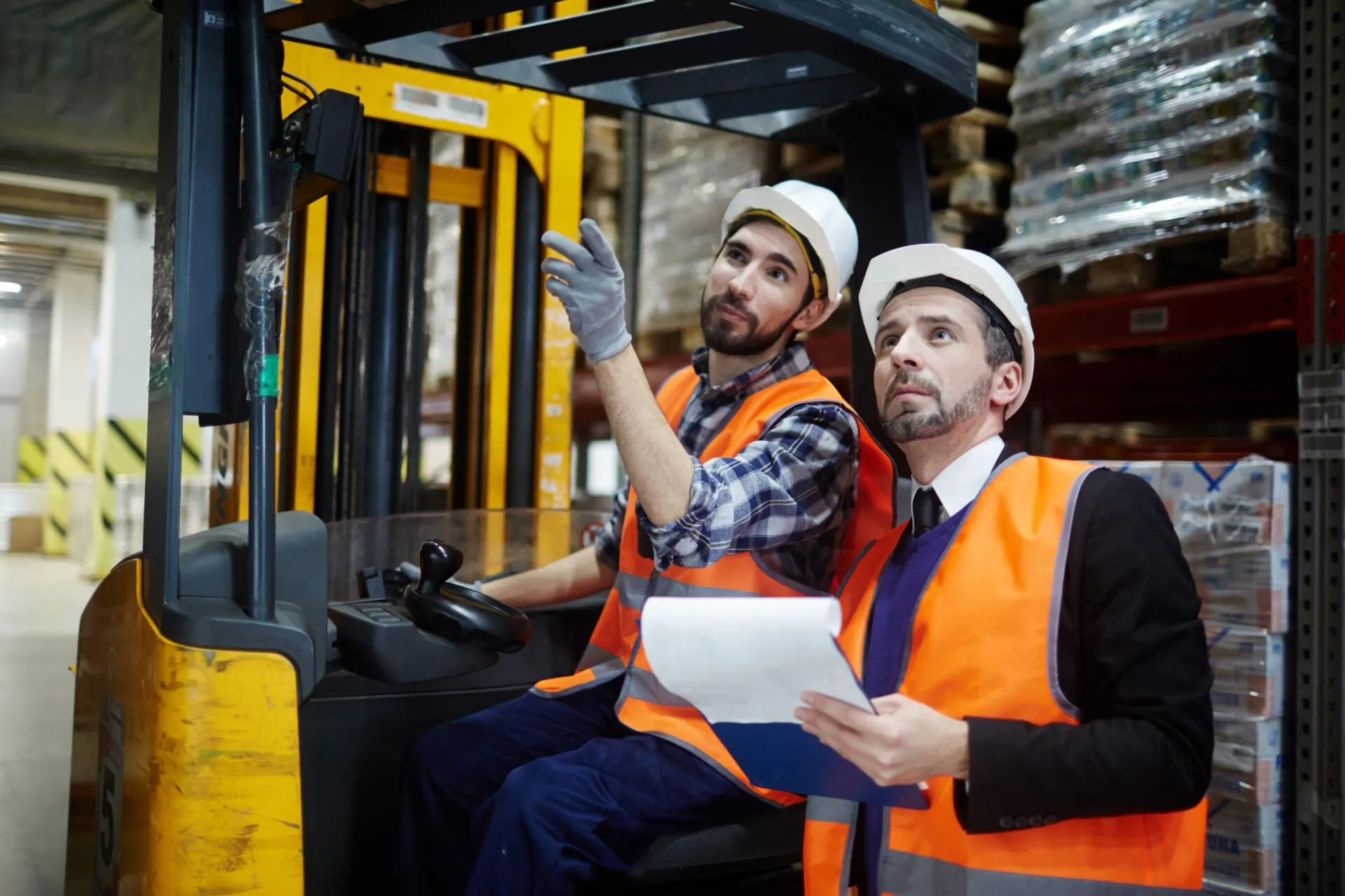  Two male warehouse workers are standing next to a yellow forklift. The man on the left is seated in the forklift, wearing a plaid shirt, a bright orange safety vest, and a white hard hat. He is looking up and to the side, pointing with a gloved hand. The man on the right, who appears to be a manager, is standing, also wearing a white hard hat and a bright orange safety vest over a dark suit jacket and tie. He is holding a clipboard and looking in the direction his colleague is pointing. The background shows tall shelves and stacked goods, indicating a busy warehouse environment.