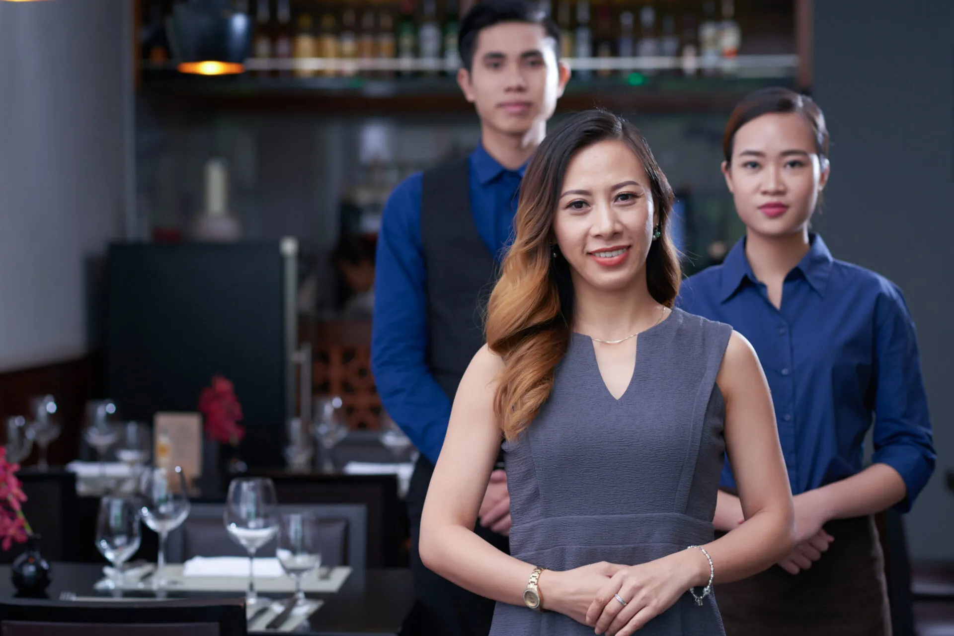 A portrait of three restaurant staff members. A woman with an ombre hairstyle and a gray sleeveless dress is in the foreground, smiling at the camera. Her hands are clasped in front of her. Behind her, a man in a blue shirt and a black vest stands on the left, and a woman in a blue shirt stands on the right. Both of them are looking straight ahead with neutral expressions. The background is a slightly blurred restaurant with a bar, a set table with wine glasses, and a dark, warm ambiance.