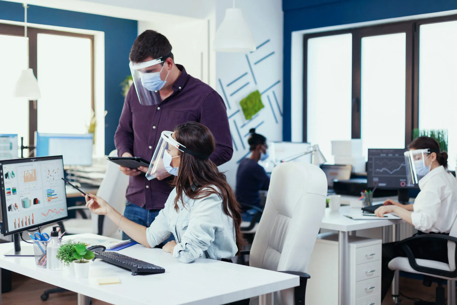 An office setting where three people are working while wearing protective masks and face shields. In the foreground, a woman seated at a white desk is pointing at a computer monitor displaying graphs and charts. A man stands beside her, looking at the monitor and holding a tablet. In the background, another woman is seated at a desk, looking at her own computer. The office has blue accent walls, and large windows, and appears bright and modern.
