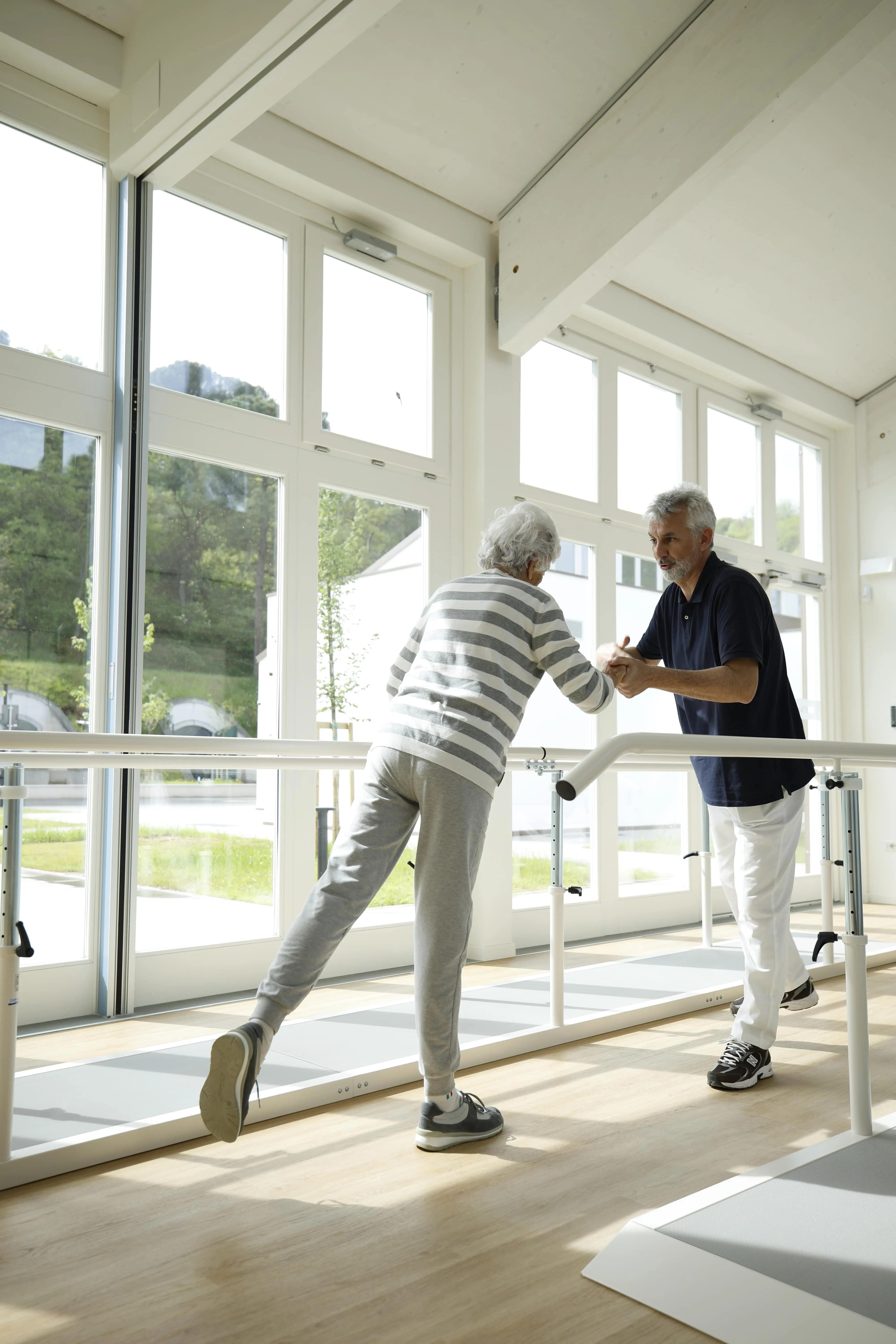 Un fisioterapista aiuta una paziente anziana a eseguire un esercizio di riabilitazione su una parallela in una sala luminosa con grandi finestre e vista su un ambiente naturale