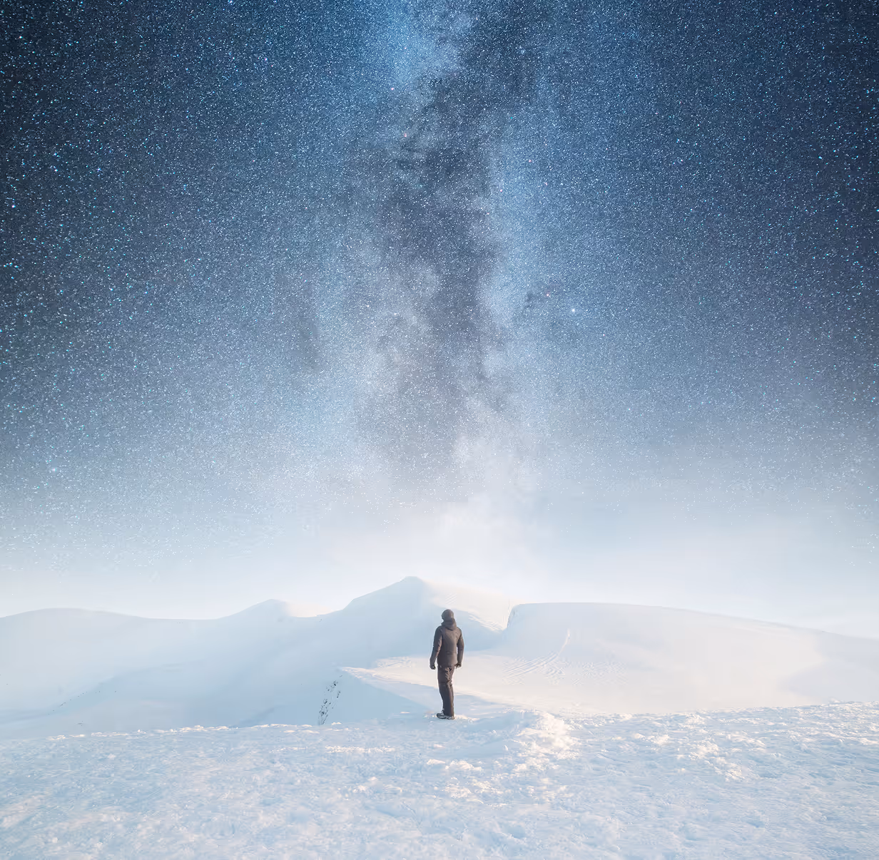 Person steht auf schneebedecktem Berg unter klarem Nachthimmel mit sichtbarer Milchstraße.