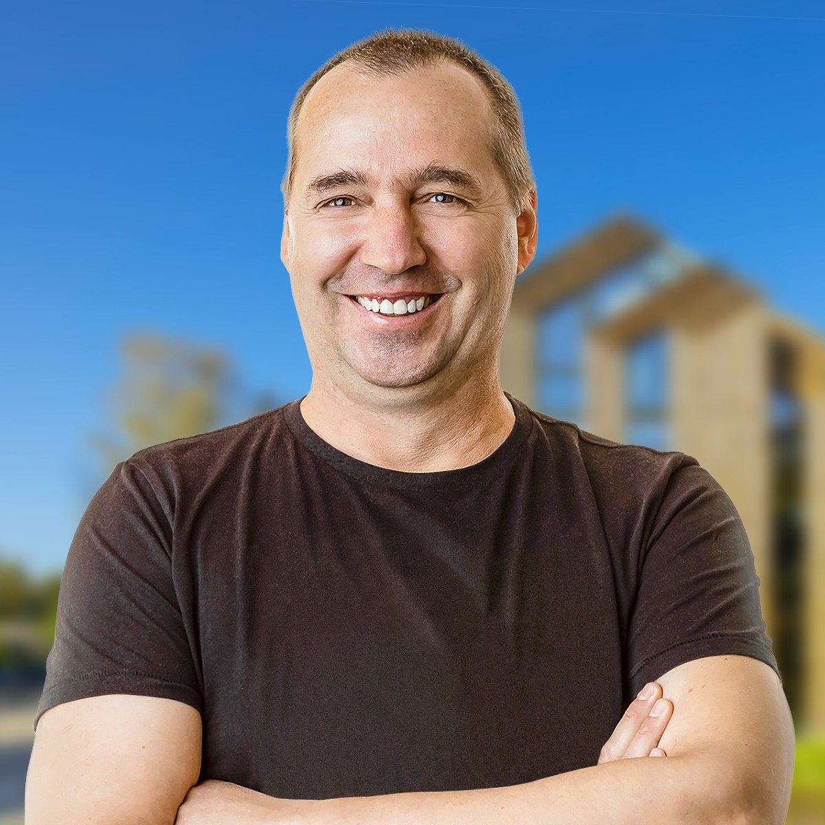 Ein Mann in einem schwarzen T-Shirt lächelt mit verschränkten Armen. Er steht im Freien, im Hintergrund sind ein verschwommenes modernes Gebäude und ein klarer blauer Himmel zu sehen.