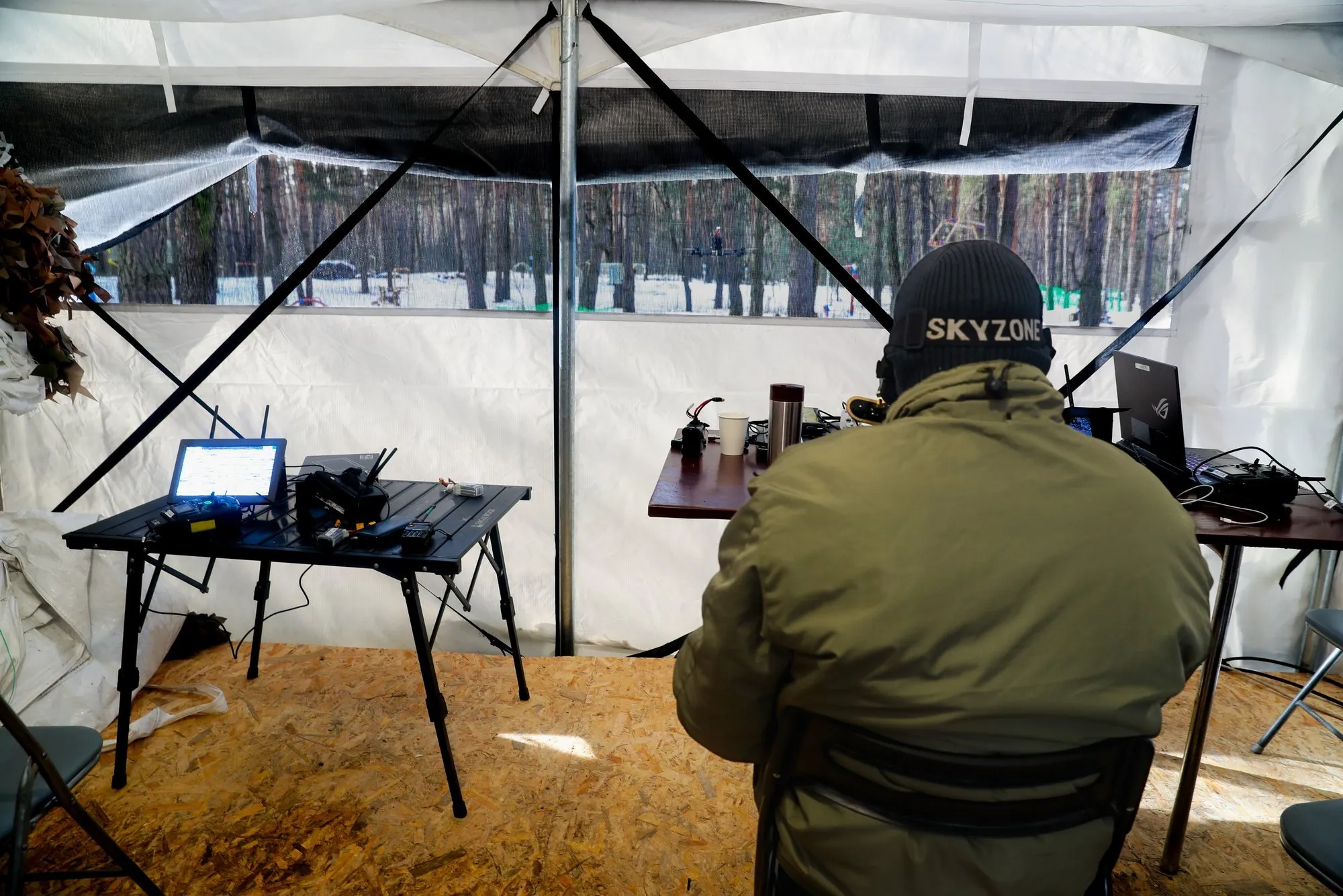 Людина в куртці та шапці зі словом SKYZONE сидить за столом із електронними пристроями у наметі з видом на зимовий ліс.