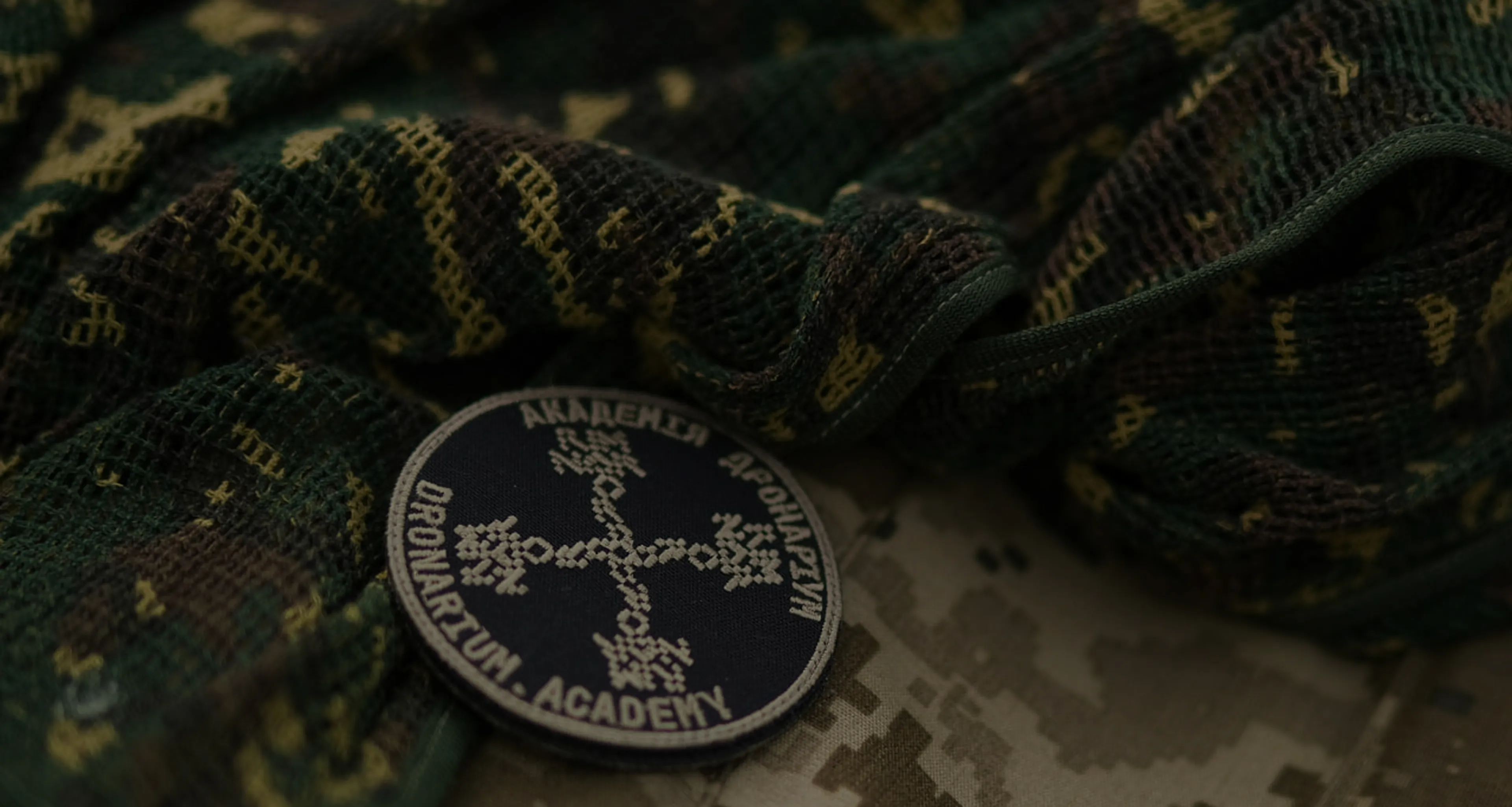Нашивка Академії дронаріум із зображенням хрестоподібного орнаменту на темно-синьому фоні лежить на камуфляжній тканині в зелених та коричневих тонах.