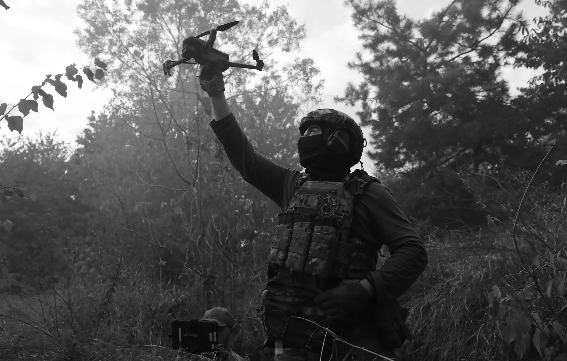 Солдат у військовій формі та тактичному спорядженні в лісі піднімає дрон у руці, готуючись до запуску.