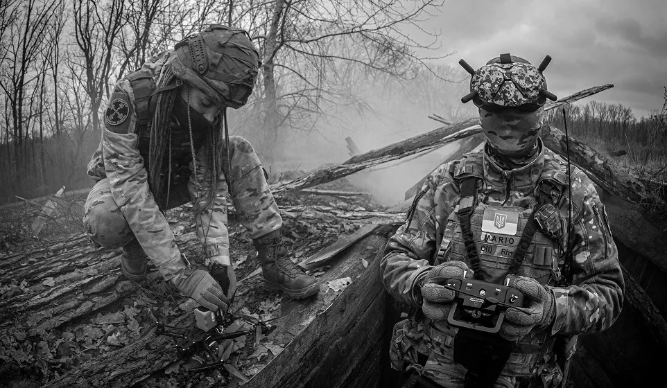 Двоє військових у камуфляжній формі на тлі лісу та зруйнованої місцевості: один налаштовує квадрокоптер, інший керує пультом дистанційного керування.