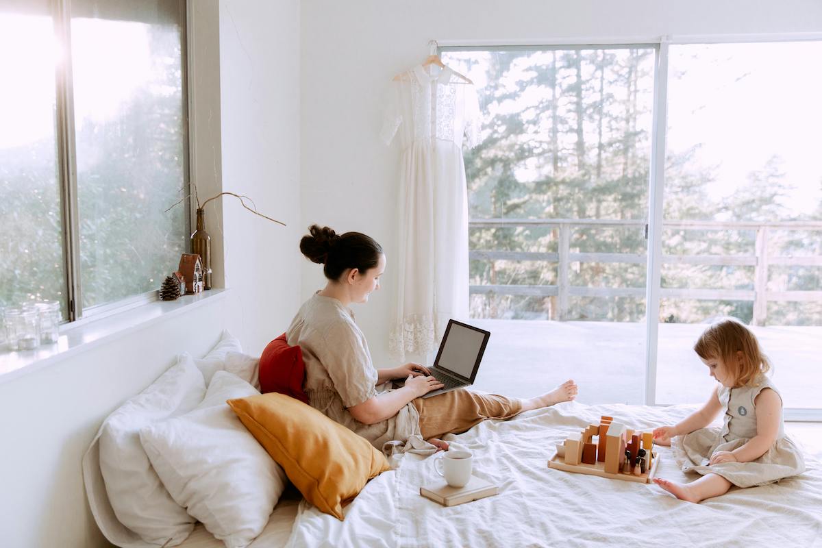 mom on her computer while child plays on the bed