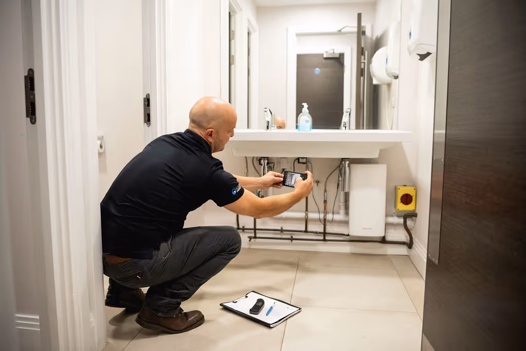 A person in a bathroom uses a smartphone to take a photo of pipes under a sink.