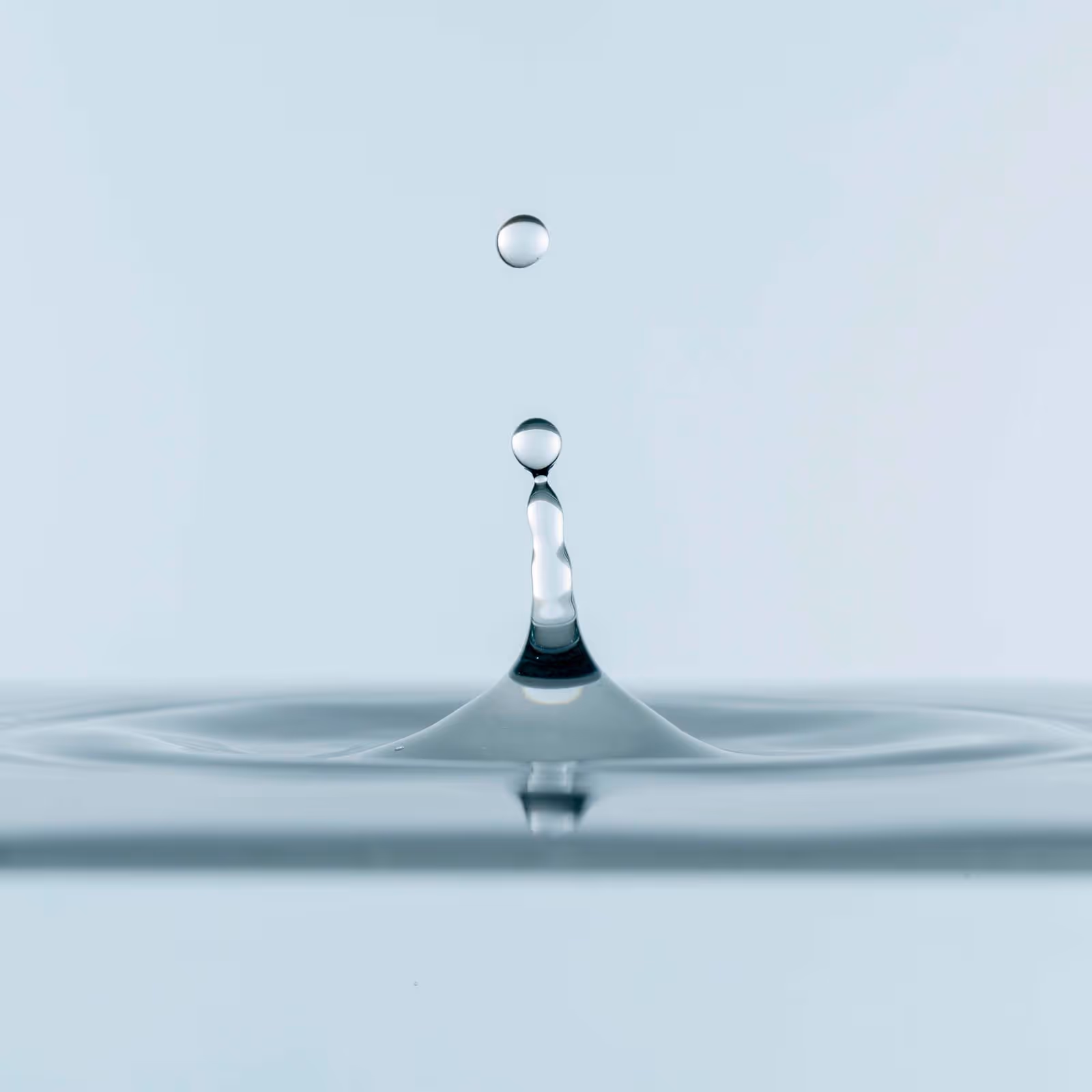 A water droplet falls and creates a splash in a calm water surface.