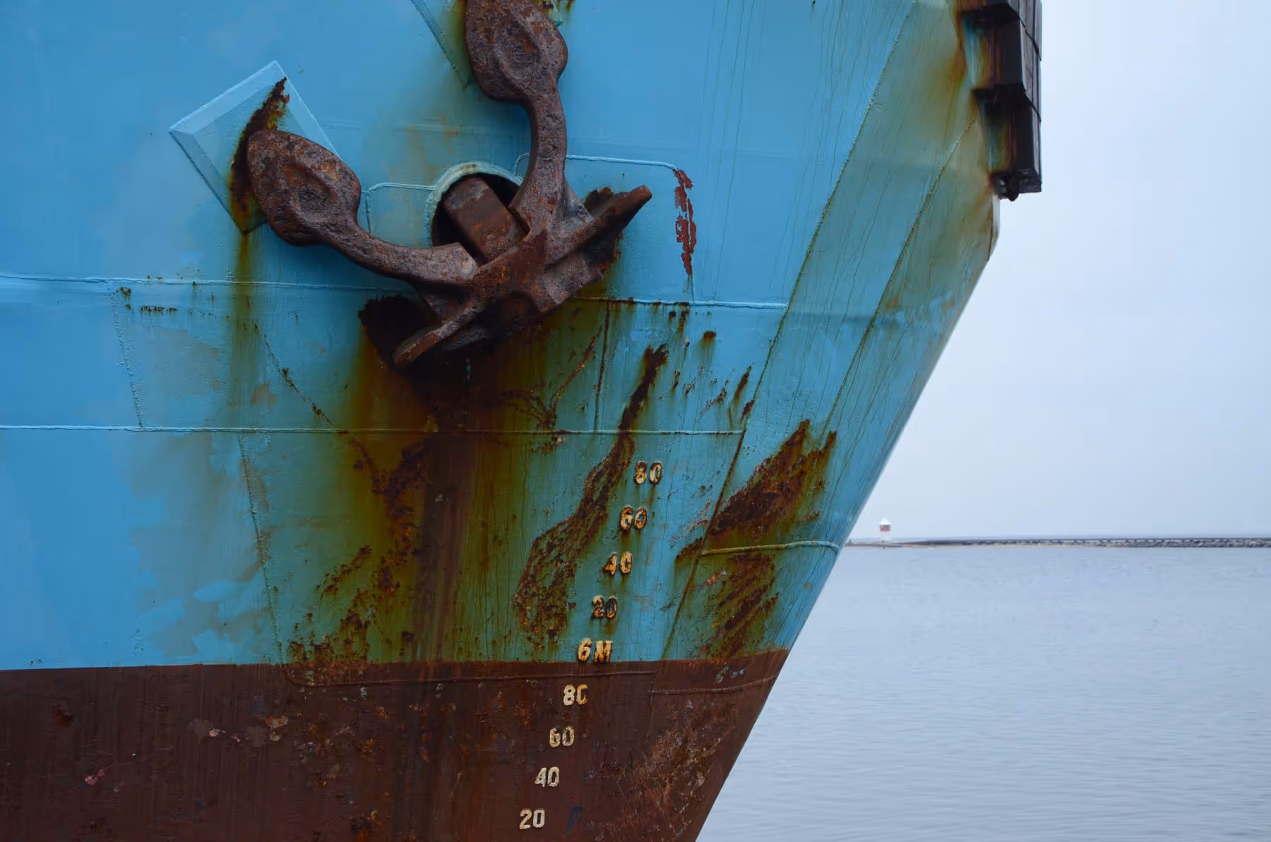 Close-up of a ship's rusted anchor and hull, showing waterline measurements in a harbor.