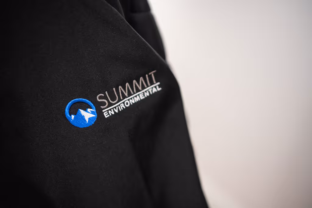 Close-up of a black jacket featuring the "Summit Environmental" logo embroidered in blue and white.