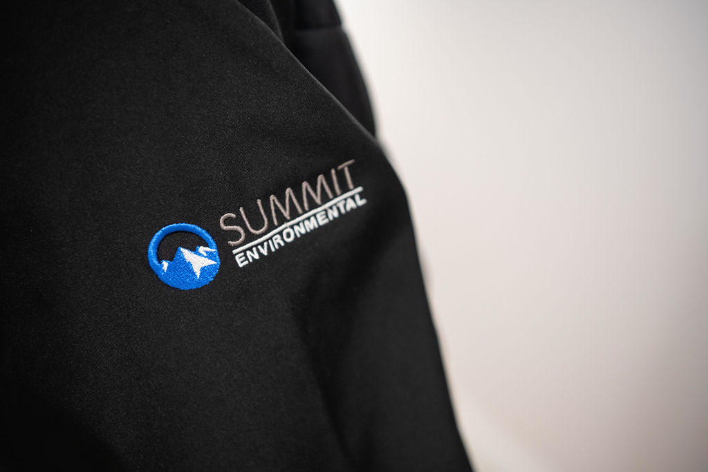 Close-up of a black jacket featuring the "Summit Environmental" logo embroidered in blue and white.