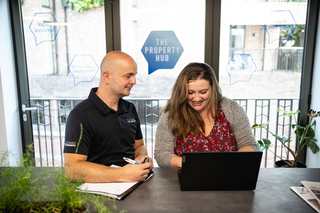 A man and woman sit at a table with a laptop, smiling and discussing something together.