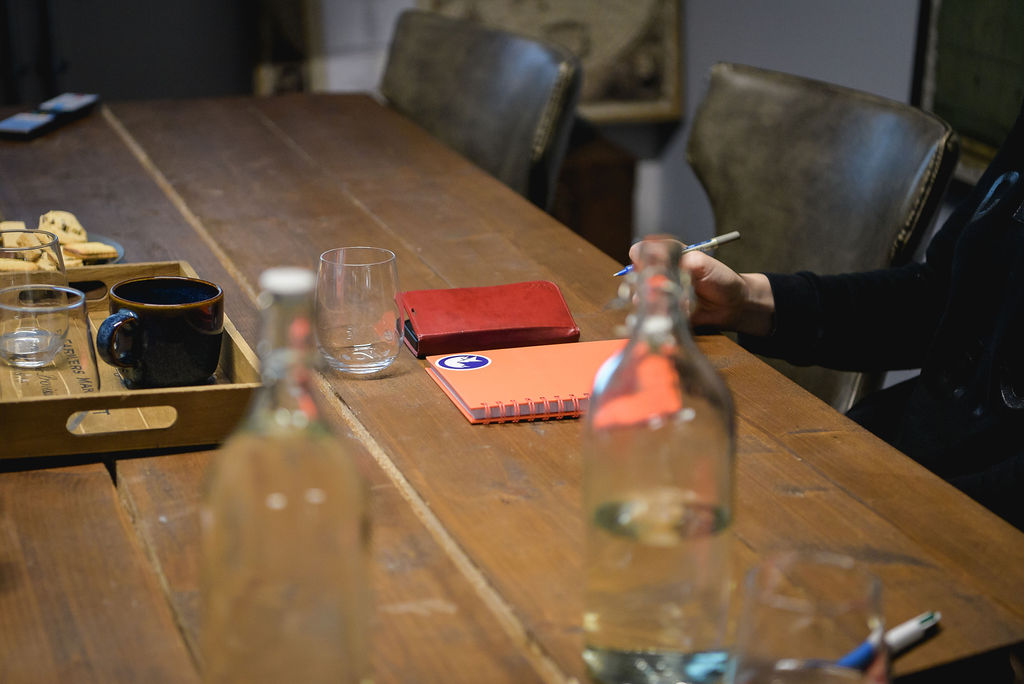 A person holding a pen sits at a table with notebooks, cups, and glass bottles.
