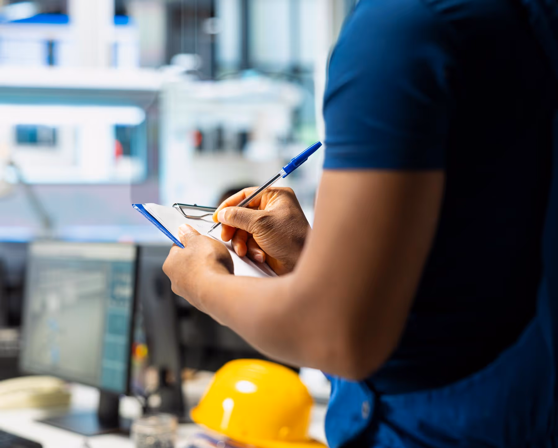 Person writing on a clipboard with a pen, inside an office. A yellow hard hat is nearby.