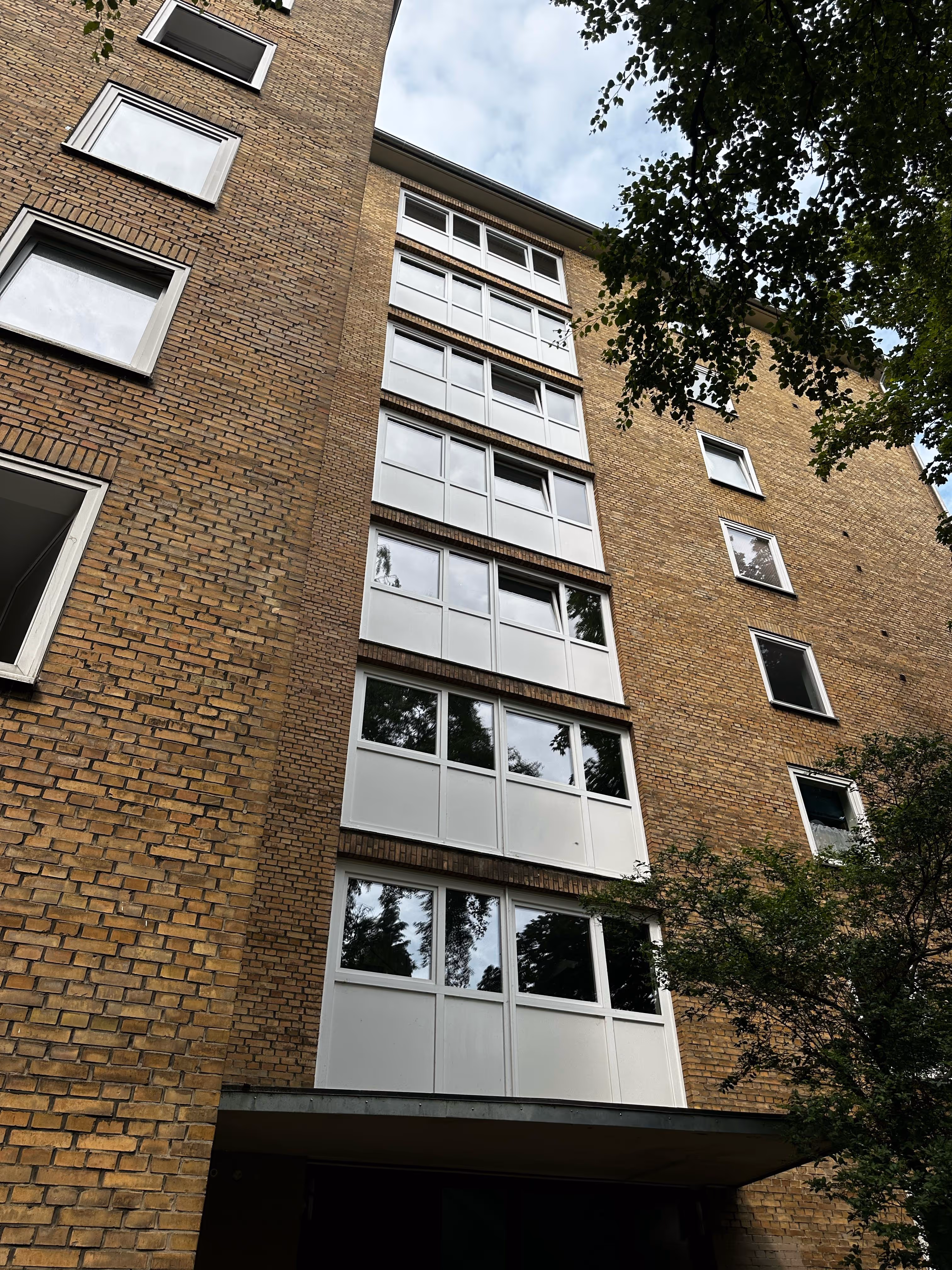 Blick nach oben auf eine hohe Backsteinwohnanlage mit weißen Fenstern, umgeben von Bäumen und bewölktem Himmel.