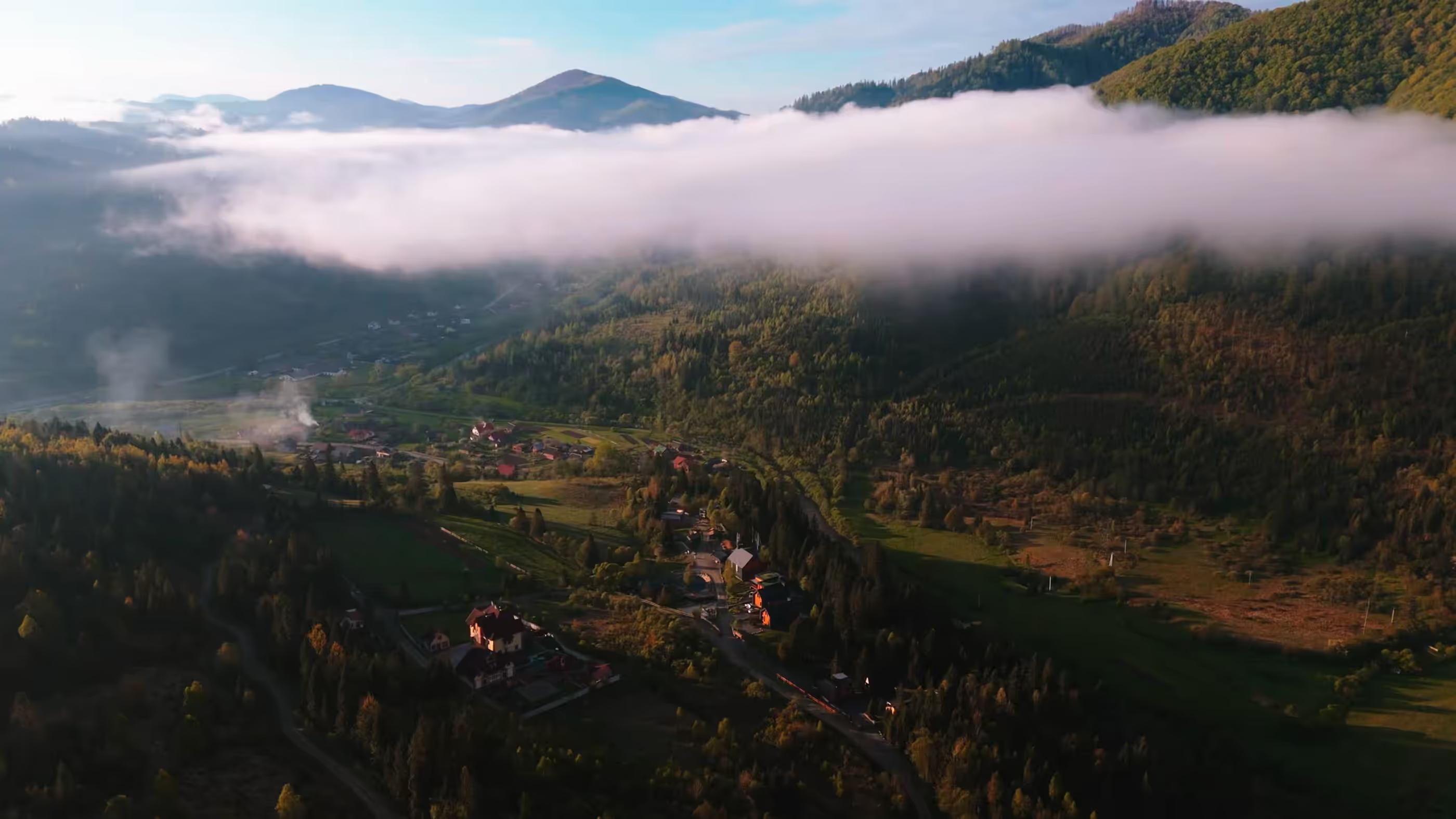 Mountain valley with houses, green trees, and a low white cloud covering part of the hillside.