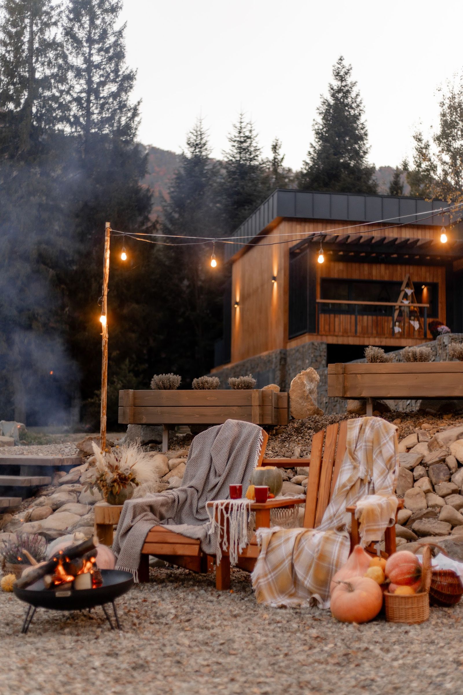 Two wooden chairs with blankets near a fire pit surrounded by pumpkins in a cozy outdoor setting.