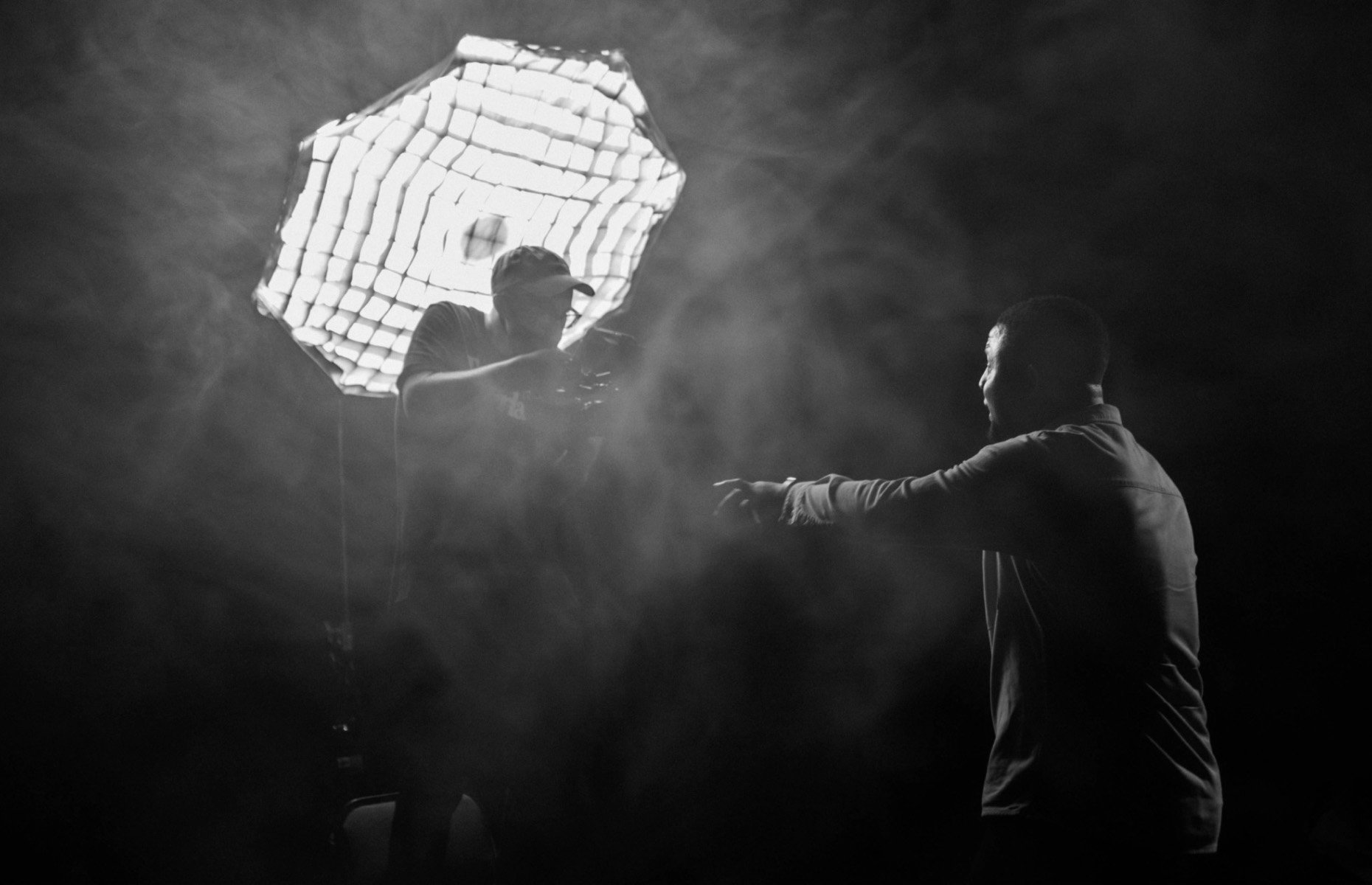 Black and white photo of a photographer adjusting a camera under a large studio light, facing a man who gestures with his hands.