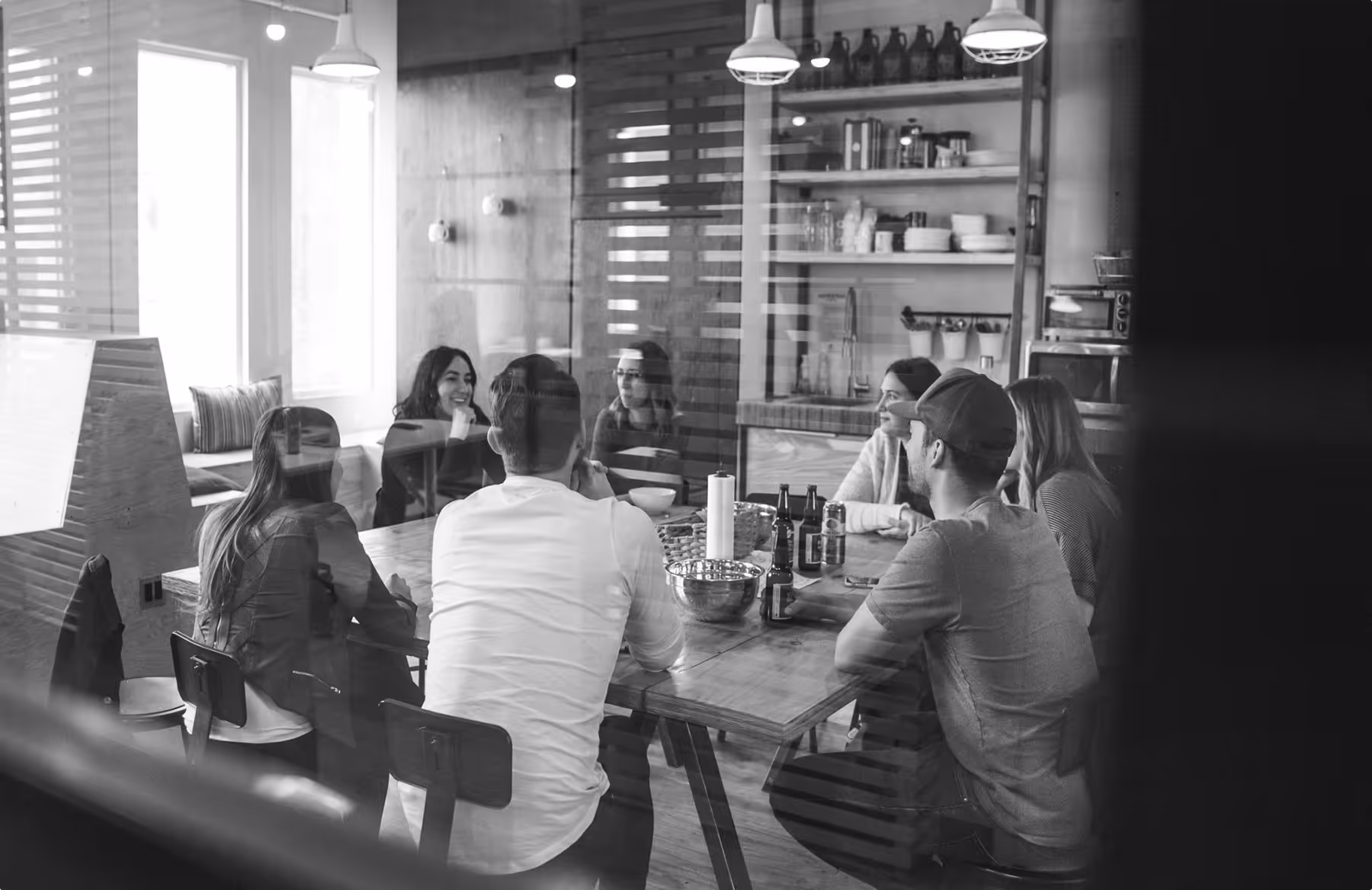 Group of seven people sitting around a table in a modern kitchen, engaged in conversation with drinks and snacks on the table.