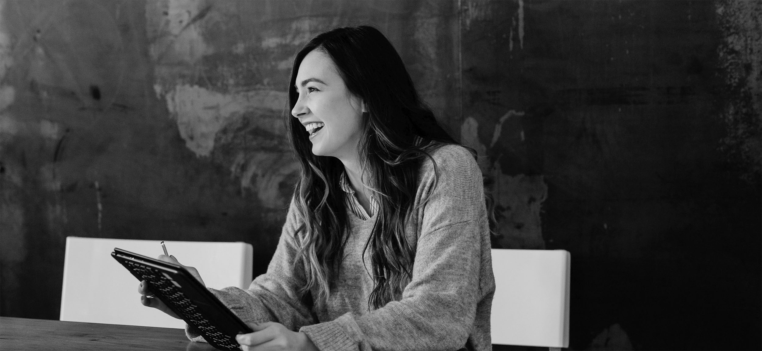 Smiling woman with long hair sitting at a table holding a tablet and stylus.