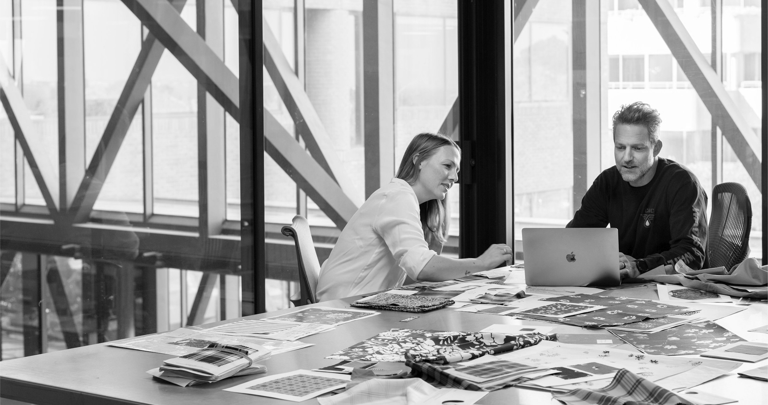 Two colleagues sitting at a table covered with fabric samples and design papers, working together on a laptop in a modern office with large glass windows.