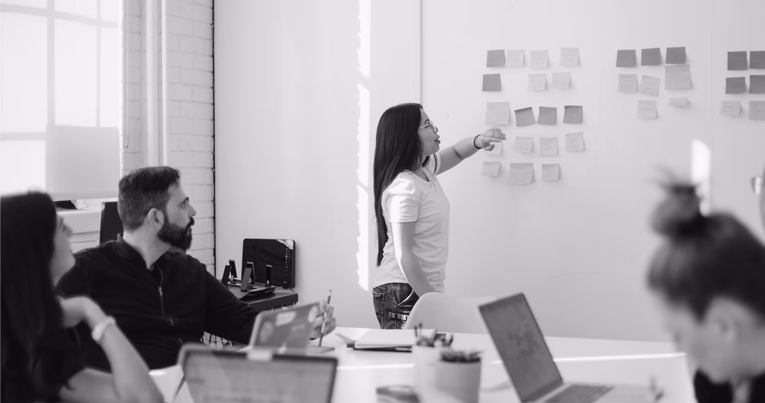 A woman points at sticky notes on a wall while colleagues watch during a meeting in a bright office.