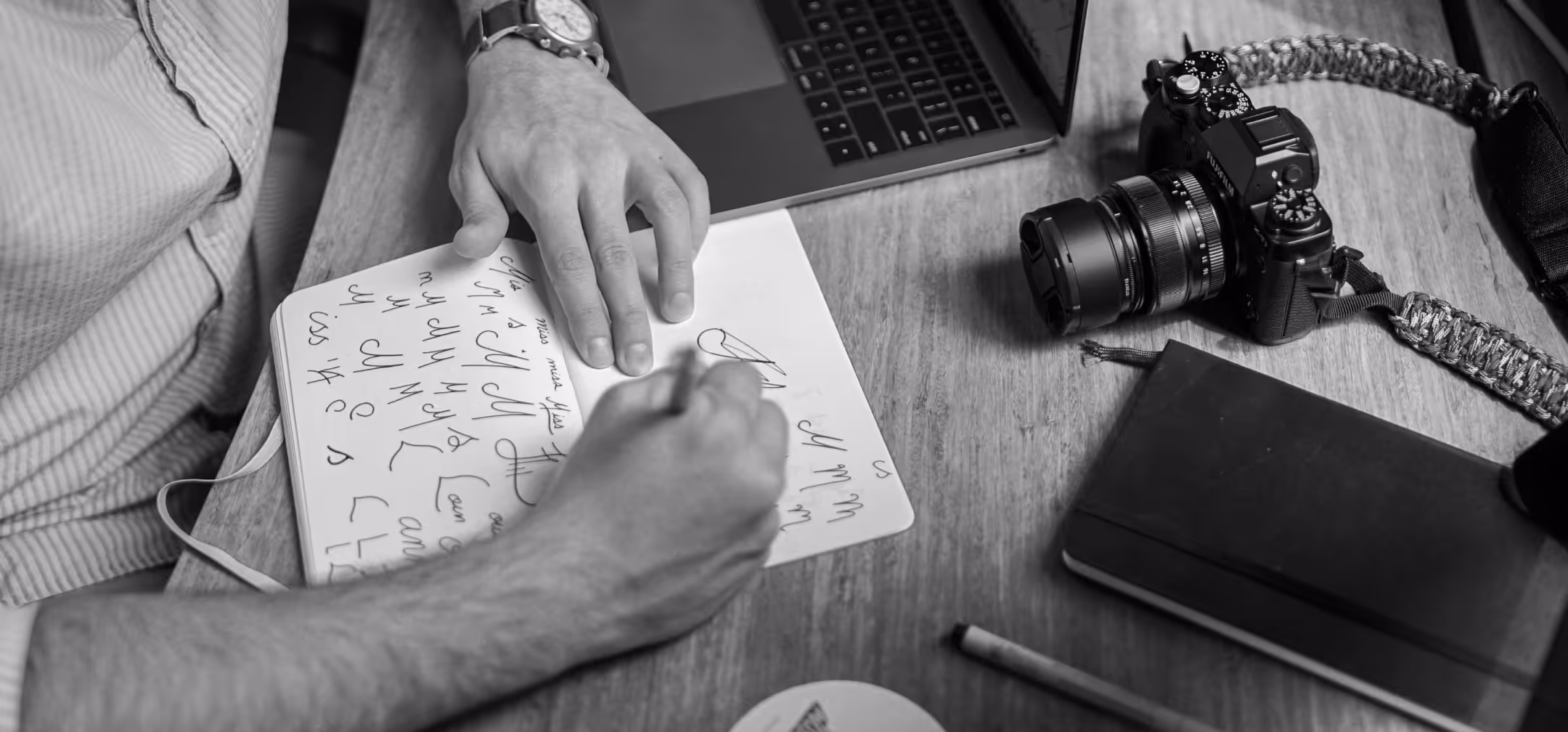Person practicing handwriting in a notebook on a wooden table with a camera, closed notebook, pencil, and laptop nearby.