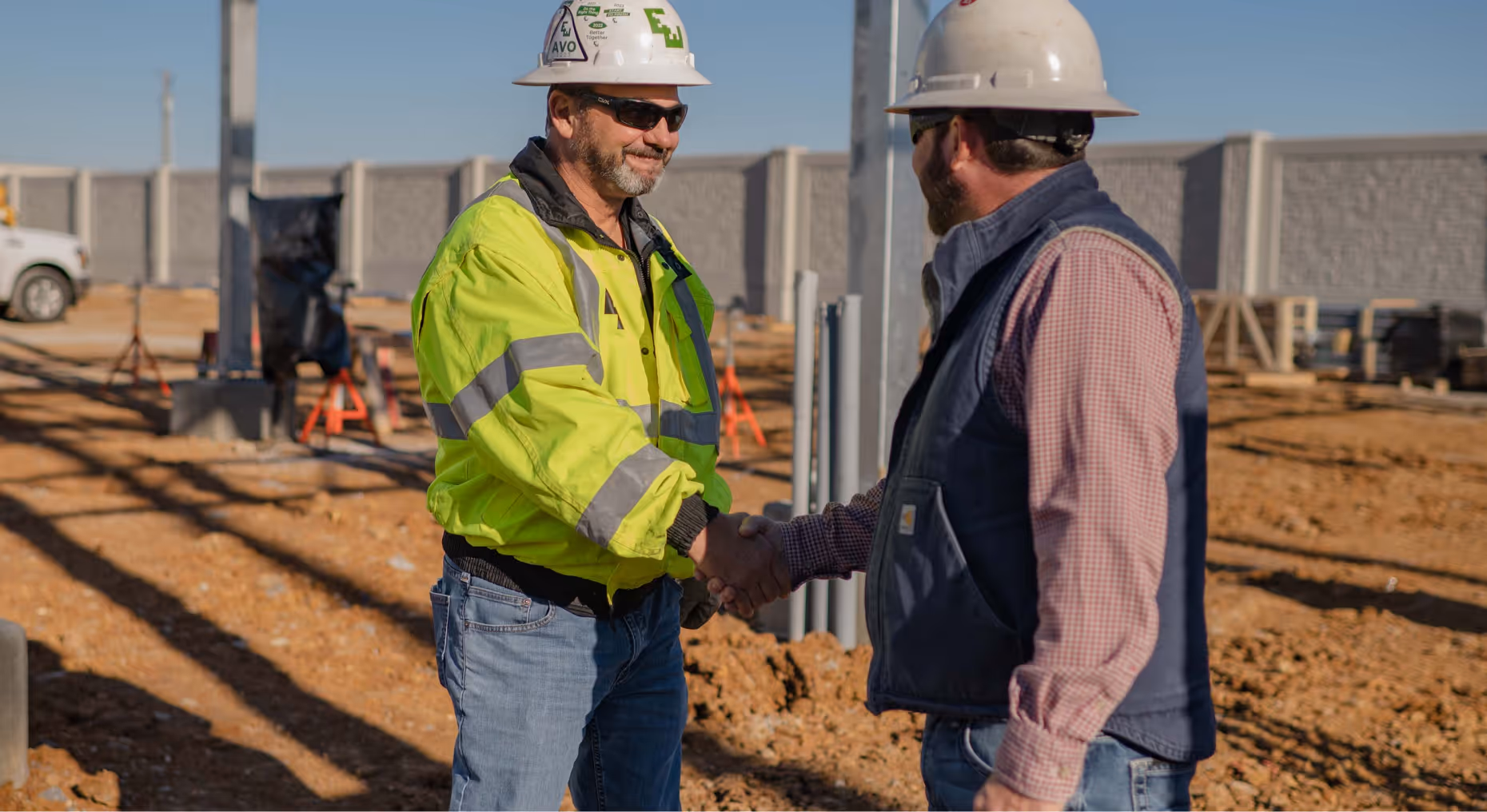 Two construction workers wearing hard hats shaking hands at a job site with dirt ground and metal poles.