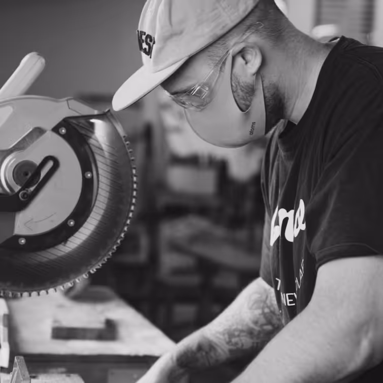 Man wearing safety glasses, a face mask, and a cap operating a circular saw in a workshop.