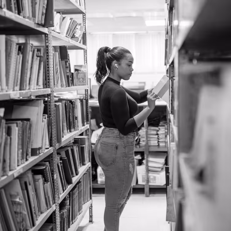 Woman in earbuds reading a book while standing between shelves in a library.
