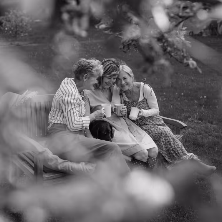 Three elderly women sitting closely on a wooden bench outdoors, smiling and holding mugs, with a dog resting on their laps.
