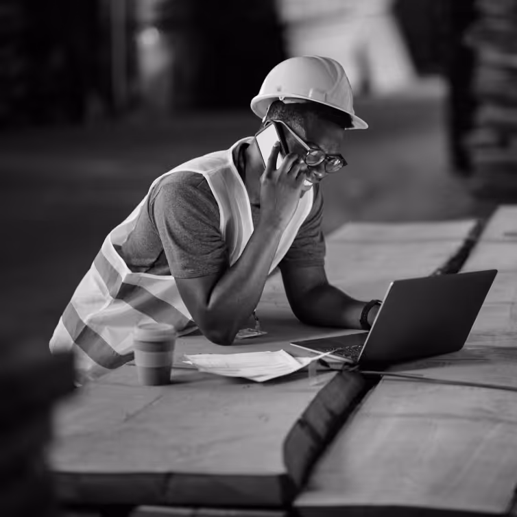 Construction worker wearing a safety vest and helmet using a laptop and talking on a phone at a wooden table.