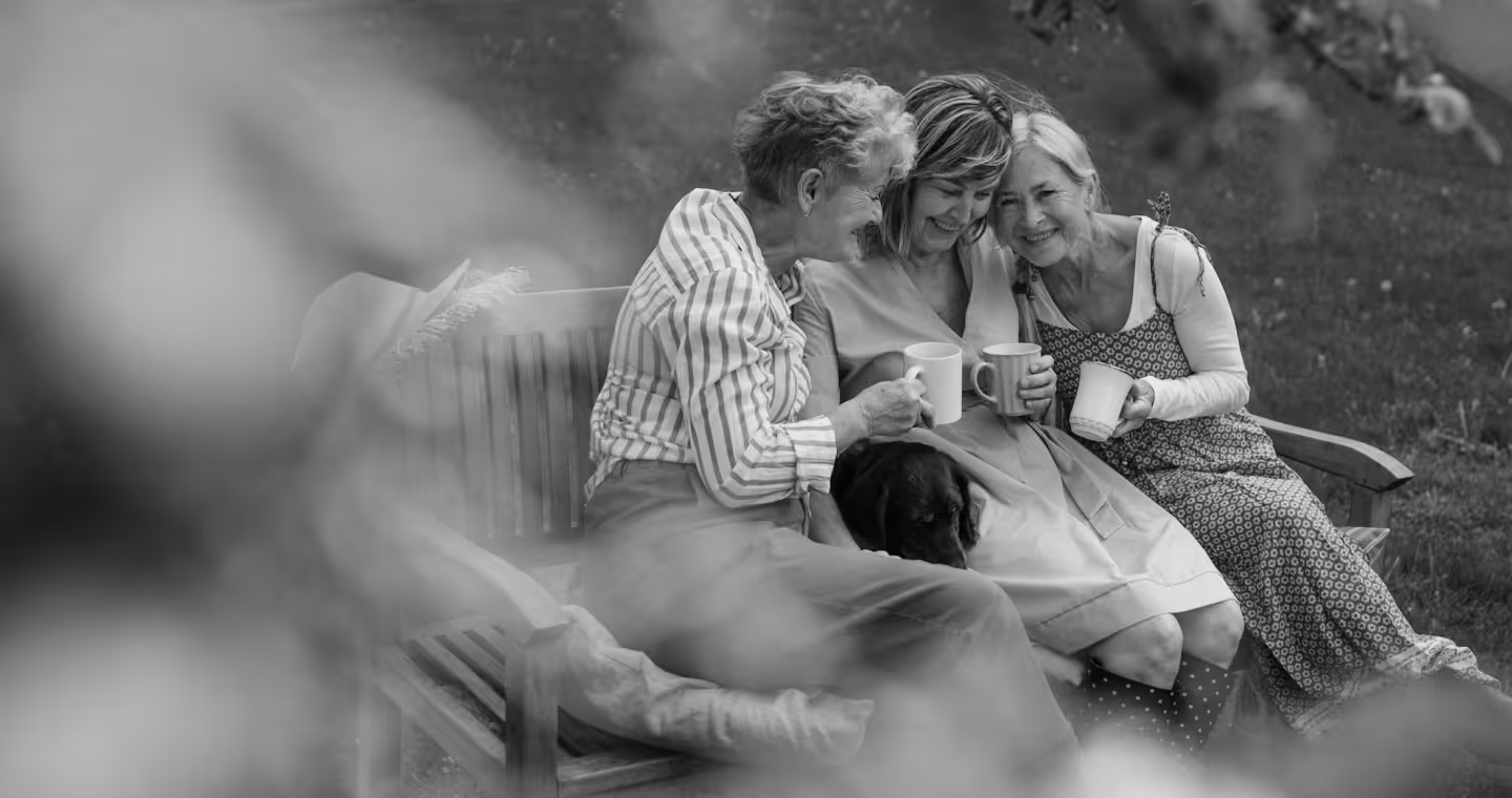 Three elderly women sitting closely on a wooden bench outdoors, smiling and holding coffee mugs, with a dog resting on their laps.
